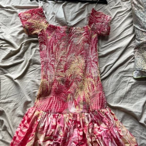 Rosa mönstrad kortklänning H&M - Supersöt kortklänning från H&M med smockad överdel och volangig kjol. Klänningen har ett tropiskt mönster i rosa, beige och gult, korta ärmar och offshoulder-modell. Perfekt för sommarens alla tillfällen!