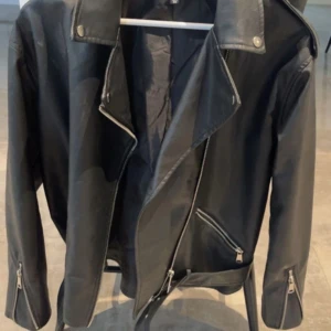 Svart skinnjacka från Missguided - Snygg svart skinnjacka från Missguided med klassisk bikerstil. Jackan har silvriga dragkedjor, bred krage med tryckknappar och bälte i midjan. Perfekt för att lyfta vilken outfit som helst och ge en edgy vibe.