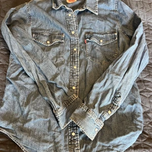 Levi's jeansskjorta med tryckknappar M - Klassisk jeansskjorta från Levi's i mellanblå denim. Skjortan har två bröstfickor med lock och tryckknappar, samt långa ärmar. Perfekt för en avslappnad och cool stil. Ikonisk röd Levi's-tagg på ena fickan.