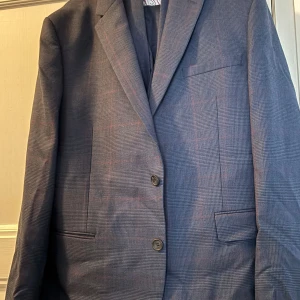 Rutig blå kavaj Ted Baker 36R - Snygg rutig kavaj från Ted Baker i mörkblått med diskreta röda linjer. Slim passform och klassisk design med två knappar framtill och flera fickor. Fodret är mönstrat i vitt och rosa. Perfekt för dig som vill sticka ut med stil.