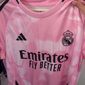 Rosa Real Madrid fotbollströja Adidas - Snygg rosa Real Madrid fotbollströja från Adidas med svart klubbmärke och sponsortryck på bröstet. Tröjan har korta ärmar och är tillverkad i ett lätt och ventilerande material, perfekt för träning eller match. Pris kan diskuteras 