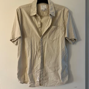 Beige kortärmad skjorta från 157 - Stilren beige skjorta från 157 i storlek M. Skjortan har korta ärmar, klassisk krage, bröstficka och knäppning framtill. Perfekt för en avslappnad look och lätt att matcha med andra plagg.