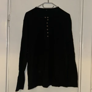 Svart långärmad tröja med knappar - Svart långärmad tröja i storlek L med rund halsringning och snygga knappar framtill. Tröjan har en enkel och clean look, perfekt för dig som gillar stilrena plagg. Materialet känns mjukt och lätt, och passformen är normal.