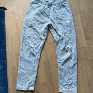 Grå mjukisbyxor från Nike - Gråa mjukisbyxor från Nike med elastisk midja och sidofickor. Byxorna har en avslappnad passform och är tillverkade i mjukt bomullsmaterial. Perfekta för chill dagar eller träning. Nike-logga broderad på vänster lår.