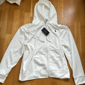 Vit hoodie från Polo Ralph Lauren - Snygg vit hoodie från Polo Ralph Lauren med dragkedja framtill och klassisk huva med snörning. Tröjan har två fickor fram och den ikoniska lilla loggan broderad på bröstet. Perfekt för en clean och stilren look. Storlek M