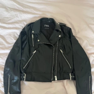 Svart skinnjacka från Zara, S - Snygg svart skinnjacka från Zara med coola dragkedjor, bälte i midjan och klassisk bikerstil. Jackan har flera fickor med dragkedja och detaljer i silver. Perfekt för dig som vill ha en edgy look och gillar statement-plagg.