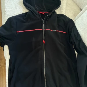 Svart hoodiejacka från Hugo med röd detalj - Svart hoodiejacka från Hugo med dragkedja och röda detaljer längs bröstet. Jackan har huva med snörning och små logotypdetaljer. Perfekt för en avslappnad och sportig stil.
