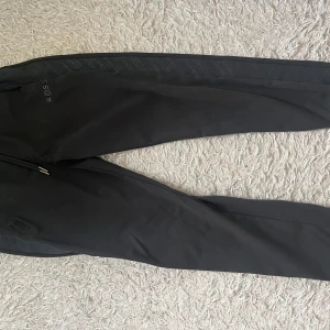 Svarta mjukisbyxor från BOSS - Svarta mjukisbyxor från BOSS med broderad logga på vänster sida. Byxorna har elastisk midja med snörning, fickor på sidorna och diskret mönstrad detalj längs benen. Perfekta för en chill och stilren look.