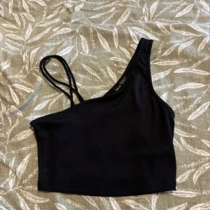 Svart oneshoulder topp från Bershka - Svart croppad topp från Bershka med asymmetrisk oneshoulder-design och dubbla smala axelband på ena sidan. Toppens material är ribbat och stretchigt, vilket ger en snygg och bekväm passform. Perfekt för dig som gillar trendiga och stilrena plagg.