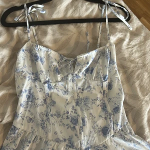 Blommig vit och blå sommarklänning - Supersöt kort klänning med vita och blå blommor, smala axelband med knyt och lätt v-ringning. Luftig och flowy modell, perfekt för varma dagar. Materialet känns som bomull eller polyester. Romantisk och fräsch  ( korsett del )  