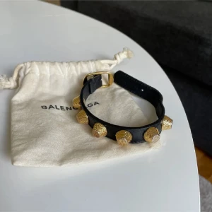 Balenciaga svart armband med guldnitar - Coolt svart armband från Balenciaga med stora, texturerade guldnitar och spänne i guld. Armbandet är gjort i skinn och har en edgy vibe som verkligen sticker ut. Perfekt accessoar för att lyfta din outfit.