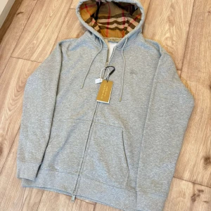 Grå hoodie från Burberry med dragkedja - Snygg grå hoodie från Burberry med klassiskt rutigt foder i huvan. Tröjan har hel dragkedja, snörning i huvan och broderad logga på bröstet. Tillverkad i mjuk bomull och har praktiska fickor framtill. Perfekt för en chill och stilren look.