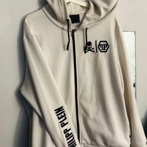 Vit hoodie från Philipp Plein - Snygg vit hoodie från Philipp Plein med svart dragkedja, huva med snörning och tryck på bröstet samt ärmen. Stora logotyper och döskalle ger en edgy vibe. Perfekt för dig som gillar streetwear och vill sticka ut. Tröjan kostar 12.500:- ny. Tänker 2000:- men priset kan diskuteras..