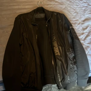 Svart bomberjacka från Plein Sport - Svart bomberjacka från Plein Sport med tryck på bröstet och ryggen. Jackan har dragkedja framtill, ribbade muddar och detaljer i blankt material. Snygg logga med tiger på både insidan och utsidan. Perfekt för dig som gillar streetwear och sportig stil. Vänstra fickdragkedjan är sönder