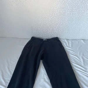 Svarta mjukisbyxor från W23 - Svarta mjukisbyxor från W23 med dragkedjeförsedda fickor och tryckt logga på benet. Byxorna har en avslappnad passform och är tillverkade i mjukt bomullsmaterial, perfekta för chill dagar eller träning.