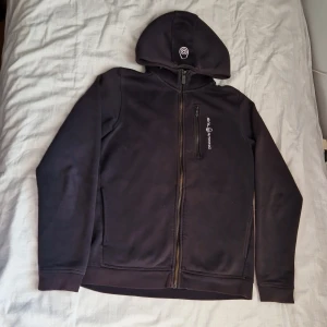 Svart hoodie från Sail Racing - Svart hoodie från Sail Racing med hel dragkedja och broderad logga på bröstet och huvan. Tröjan har lång ärm, huva och en bröstficka med dragkedja. Perfekt för dig som gillar stilren och sportig look. 