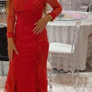 Röd långklänning med spets och pärlor - Säljer en röd långklänning med transparenta långärmar, dekorerad med spetsapplikationer och pärlor upptill. Klänningen har figurnära passform och eleganta detaljer längs ärmslut och överdel. Perfekt för bal eller festliga tillfällen.