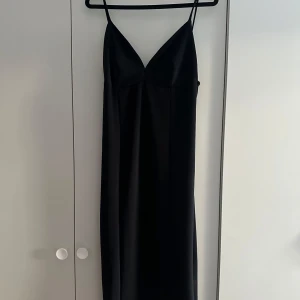 Svart klänning med smala axelband - En stilren svart långklänning med smala axelband och v-ringad urringning. Klänningen har en liten slits på ens sida. som ger en elegant känsla och faller snyggt längs kroppen. Perfekt för dig som gillar minimalistisk och tidlös stil