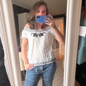 Vit offshoulder blus med broderi - Säljer en vit offshoulder blus med svart broderat blommönster framtill. Blusen har smockad kant upptill och nertill samt korta ärmar med broderad detalj.