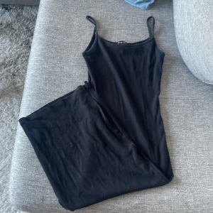 Svart långklänning från Amisu - En stilren svart långklänning från Amisu med smala axelband och enkel, rak passform och slits. Perfekt för dig som gillar minimalistisk stil och vill ha en klänning som är lätt att matcha med accessoarer. Mjuk och skön mot huden.