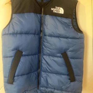 Blå dunväst från The North Face M - Säljer en blå och svart dunväst från The North Face i storlek M. Västen har quiltad design, dragkedja framtill och två snedställda fickor med svarta detaljer. Perfekt för lager-på-lager och outdoor-vibe.