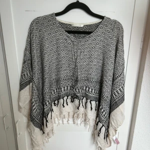 Mönstrad poncho från Pieces - Svartvit poncho från Pieces med grafiskt mönster och fransar längs nederkanten. Oversized och loose fit, perfekt att slänga på sig över en enkel outfit. Tjockare bomullsmaterial som känns både mjukt och svalt. Snygg detalj med svarta tofsar framtill.