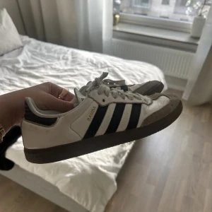 Adidas Samba vita och svarta sneakers - Klassiska Adidas Samba sneakers i vitt skinn med svarta ränder och grå mockadetaljer vid tån. Låg modell med brun gummisula och snörning. Perfekta för en sportig och stilren look. Ser lite smutsiga ut, men går att fixa med att tvätta av dem samt tvätta snörena!!
