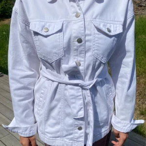 Vit jeansjacka med bälte - Snygg vit jeansjacka med bälte i midjan och stora bröstfickor med knappar. Jackan har långa ärmar, klassisk krage och knäppning framtill med silvriga tryckknappar. Perfekt för dig som vill ha en fräsch och trendig look.