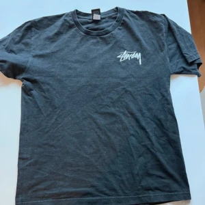 Svart Stüssy t-shirt retro - Svart t-shirt från Stüssy med vit logga på bröstet och stort skateboardinspirerat tryck på ryggen. Klassisk passform med rund hals och korta ärmar. Perfekt för dig som gillar streetwear och skatekultur.
