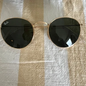 Ray-Ban runda solglasögon guld - Snygga Ray-Ban solglasögon med rund form och tunna guldfärgade metallbågar. Mörka linser och klassisk logga i hörnet. Kommer med svart fodral och putsduk. Perfekta för att lyfta din stil i solen.