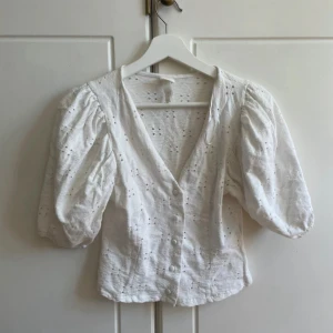 Vit blus med puffärm från H&M - Supersöt vit blus från H&M med broderade hål, puffiga korta ärmar och v-ringning. Blusen har knappar framtill och är croppad i modellen. Perfekt för en fräsch och somrig look. I väldigt bra skick❤️