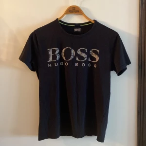 Svart BOSS t-shirt med logga - Svart t-shirt från Hugo Boss i storlek L. Klassisk modell med rund hals och stor BOSS-logga framtill i grått och vitt. Tillverkad i mjuk bomull som känns skön mot huden. Perfekt för dig som gillar stilrena och snygga plagg med tydlig branding.