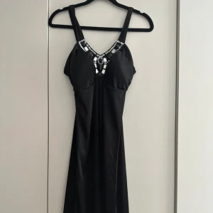 Svart långklänning med strassdetaljer - Elegant svart långklänning med breda axelband och v-ringning dekorerad med glittrande strass och paljetter. Klänningen har en mjuk, följsam passform och är perfekt för en snygg och glamorös look. Materialet är glansigt och känns lyxigt mot huden