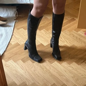 Svarta skinnstövlar – italienskt läder - Svarta stövlar i skinn med hög klack och fyrkantig tå. Klassisk modell med dragkedja på insidan och platt sula framtill samt markerad klack. Strl 40.