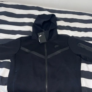Svart Nike tech kofta - Säljer en svart hoodiejacka från Nike med dragkedja och huva. Jackan har en diskret Nike-logga på bröstet och en praktisk ficka med dragkedja på ärmen. Lite mer loose fit. Storlek M.