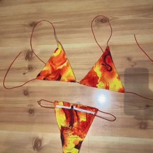 Färgstark bikini från PrettyLittleThing - Snygg bikini i triangelmodell från PrettyLittleThing med unikt mönster i eldiga nyanser av orange, rött och gult. Underdel har stretchiga knytband. Ena bandet är lite trasigt på ena sidan men sitter fortfarande starkt ihop och håller ej på att gå sönder så som det ser ut. Perfekt för stranden eller poolen när du vill sticka ut. Passar S/M.
