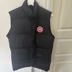 Svart dunväst från Canada Goose M - Svart dunväst från Canada Goose med klassisk logga på bröstet och broderad etikett på ryggen. Västen har hög krage, två stora fickor framtill och stängs med både dragkedja och knappar. Perfekt för kalla dagar och riktigt snygg streetstil.
