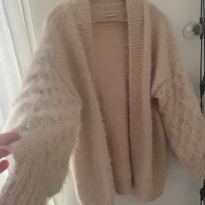Beige/Rosa mjuk stickad kofta från d'NADA - Supermjuk och oversized beige stickad kofta från d'NADA. Koftan har breda ribbade muddar, fluffiga ärmar med strukturstickat mönster och öppen front. Perfekt för lager-på-lager och chill vibes under kyliga dagar.