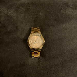 Guldig klocka från Guess - Stilren guldig klocka från Guess med metallarmband och rund urtavla. Klockan har flera små urtavlor och tydliga siffror, perfekt för dig som vill ha en snygg accessoar som sticker ut. Länken är i metall och har ett klassiskt spänne med Guess-logga.