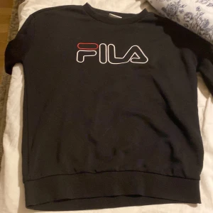 Svart sweatshirt från FILA - Svart sweatshirt från FILA med stor logga i vitt och rött framtill. Klassisk rund halsringning och ribbade muddar vid ärmslut och nederkant. Perfekt för en sportig och avslappnad stil.