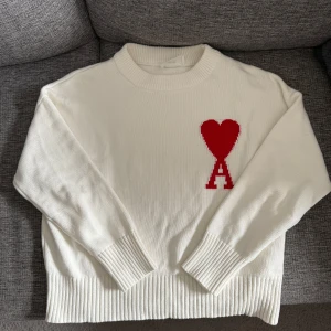 Vit stickad tröja från Ami Paris - Vit stickad tröja från Ami Paris med röd hjärtlogga och bokstaven A på bröstet. Tröjan har rund hals, ribbade muddar och är långärmad. Perfekt för dig som gillar stilrena plagg med en twist.