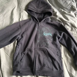 Svart hoodiejacka från Quiksilver - Svart hoodiejacka från Quiksilver med dragkedja, huva och kängurufickor. Framsidan har ett coolt tryck och broderad Quiksilver-logga i turkost. Jackan är i mjukt bomullsmaterial och har en avslappnad passform.