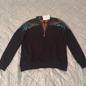 Svart tröja Marcelo Burlon med vingar - Svart sweatshirt från Marcelo Burlon med ikoniskt vingtryck i orange och turkos över axlar och hals. Tröjan har rund hals, långa ärmar och ribbade muddar. Perfekt statement piece för dig som vill sticka ut med cool design.