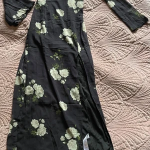 Svart blommig långklänning Boohoo - Supersnygg svart långklänning från Boohoo med vita blommor, v-ringning och långa ärmar. Klänningen har en slits på sidan och dragkedja i ryggen. Perfekt för dig som vill sticka ut med en romantisk och trendig look.