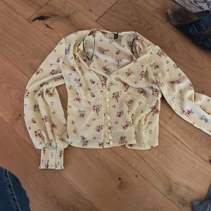 Blommig blus från H&M i ljusgul - Supersöt blommig blus från H&M i ljusgul med små rosa och röda blommor. Blusen har långa ärmar med breda smockade muddar, v-ringning och knappar framtill. Perfekt för en romantisk look och passar till både jeans och kjol.