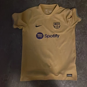 Barcelona Nike Dri-FIT fotbollströja XL - Säljer en Barcelona fotbollströja i guldbeige från Nike med Dri-FIT-material. Tröjan har korta ärmar, klubbmärke och Nike-logga på bröstet samt Spotify som sponsor. Snygga detaljer i blått och diskret mönster i tyget. Perfekt för dig som älskar Barca!