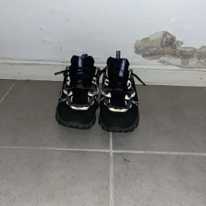 Nike React Element 55 svarta sneakers - Svarta Nike React Element 55 sneakers med vita detaljer och blå inslag på hälen. Skorna har en chunky sula med vågigt mönster, snörning och mesh-överdel för extra andningsförmåga. Swoosh-logga på sidan och reflexdetaljer för en sportig look.