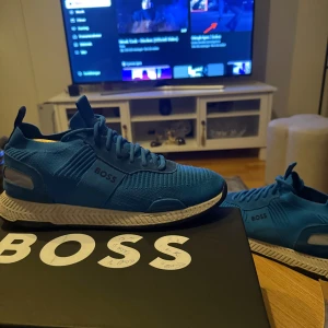 Blå sneakers från BOSS - Snygga blå sneakers från BOSS med stickad ovandel och vita detaljer på sulan. Skorna har snörning, rund tå och en sportig vibe. Materialet är en mix av textil och syntet, perfekt för dig som gillar stilrena och bekväma sneakers med modern design.