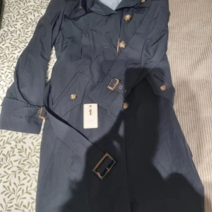 Mörkblå trenchcoat med bälte - Snygg mörkblå trenchcoat med klassiska knappar och bälte i midjan. Jackan har långa ärmar, krage och snygga spännen vid ärmslut och midja. Perfekt för dig som vill ha en stilren och tidlös look.