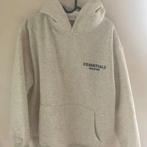Grå Essentials Fear of God hoodie - Snygg ljusgrå hoodie från Essentials Fear of God med svart logga på bröstet och ryggen. Oversized passform, stor huva och klassisk känguruficka framtill. Perfekt för en chill och trendig streetwear-look.